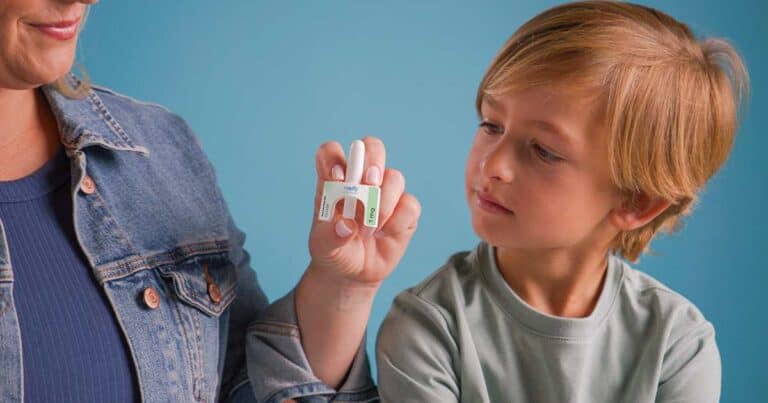 A child attentively looks at a small device being held by an adult's hand. The adult is wearing a denim jacket, and the child has short brown hair and a light green shirt. The background is a solid light blue.