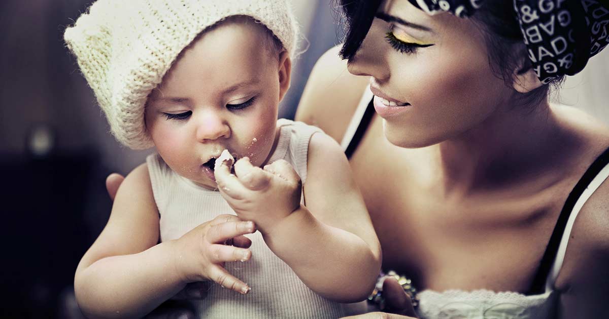 A mother with dark hair smiles at her baby who is wearing a knitted hat and a white tank top. The baby is focused on eating a piece of food, holding it with both hands. The scene is warmly lit, highlighting their joyful interaction.