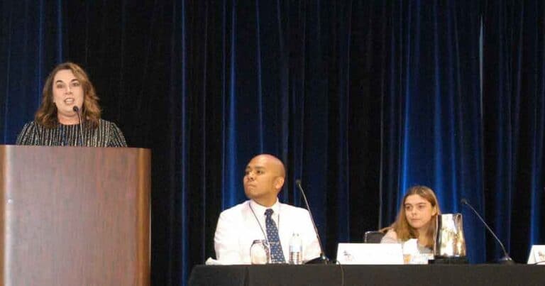 A woman speaks at a podium on a stage. Two people are seated at a table next to her, one man and one woman, both attentively listening. The background is a dark curtain.