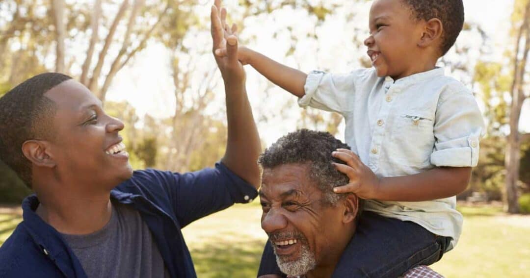 Three people outdoors: a child on an older man's shoulders, both smiling. The child is reaching out to high-five another smiling man. They are in a park with trees in the background on a sunny day.