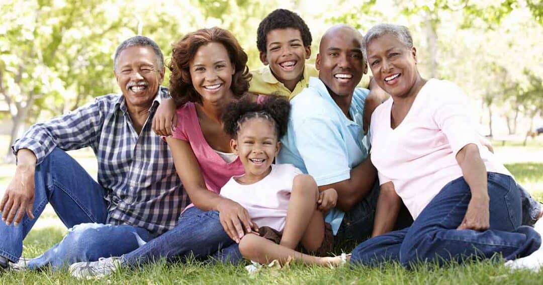 A multi-generational family of six, including three adults and three children, sits together on grass in a park. They are smiling and dressed casually. Lush green trees are visible in the background, suggesting a sunny day.