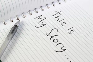 A spiral notebook with lined pages is open. A pen is resting on the paper. The handwritten text reads, "This is My Story...