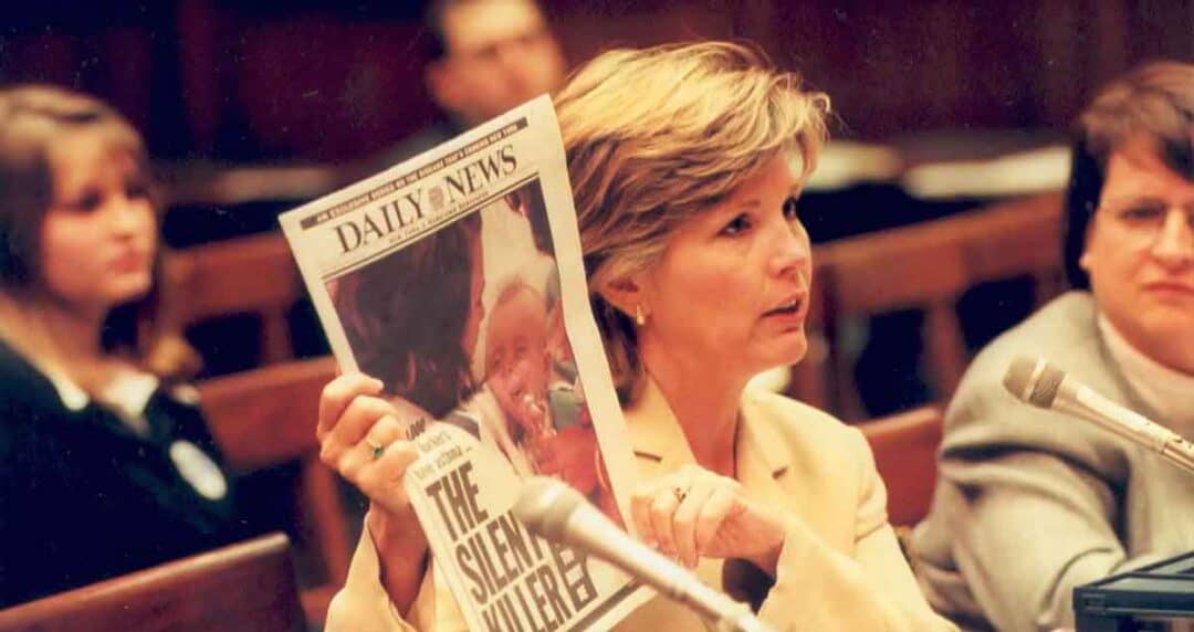 Nancy, dressed in a light-colored suit, speaks at a microphone while holding up a "Daily News" newspaper with the headline "The Silent Killer" in a formal meeting room, with others seated nearby.