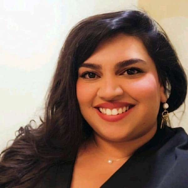 Smiling person with long dark hair and wearing a black top, pearl earrings, and a necklace. Neutral background.