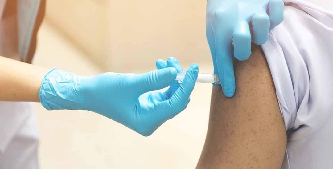 A healthcare professional wearing blue gloves administers a vaccine injection into a person's upper arm. The background is neutral, focusing on the procedure.