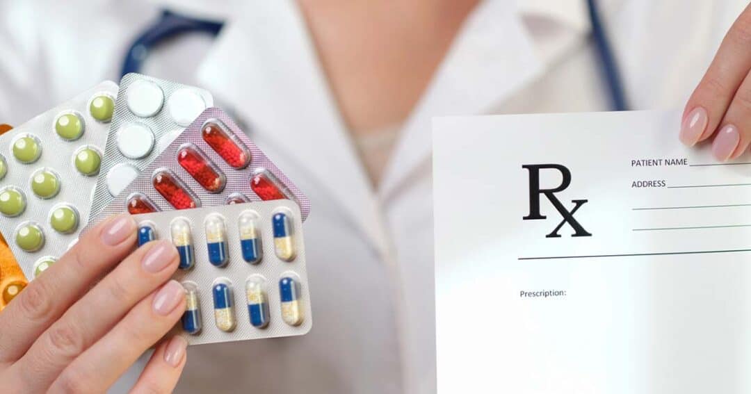 A person in a white coat holds blister packs of various pills in one hand and a blank prescription form in the other, suggesting a medical or pharmaceutical context.