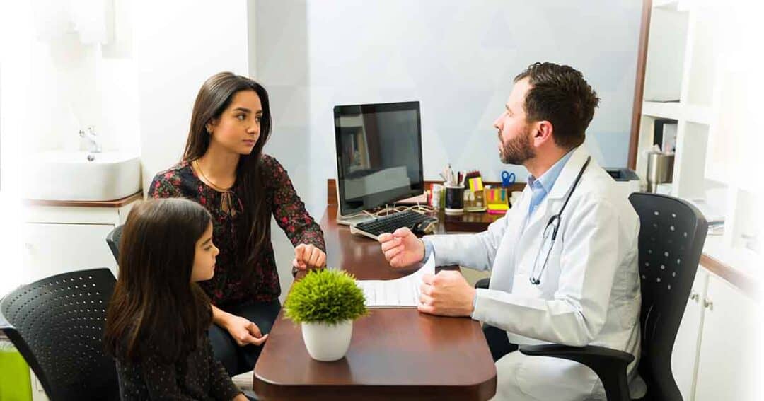 A doctor in a white coat sits at a desk talking to a woman and a young girl. The woman is sitting on a chair, and the girl is sitting next to her. An office setup with a computer and plant is in the background.