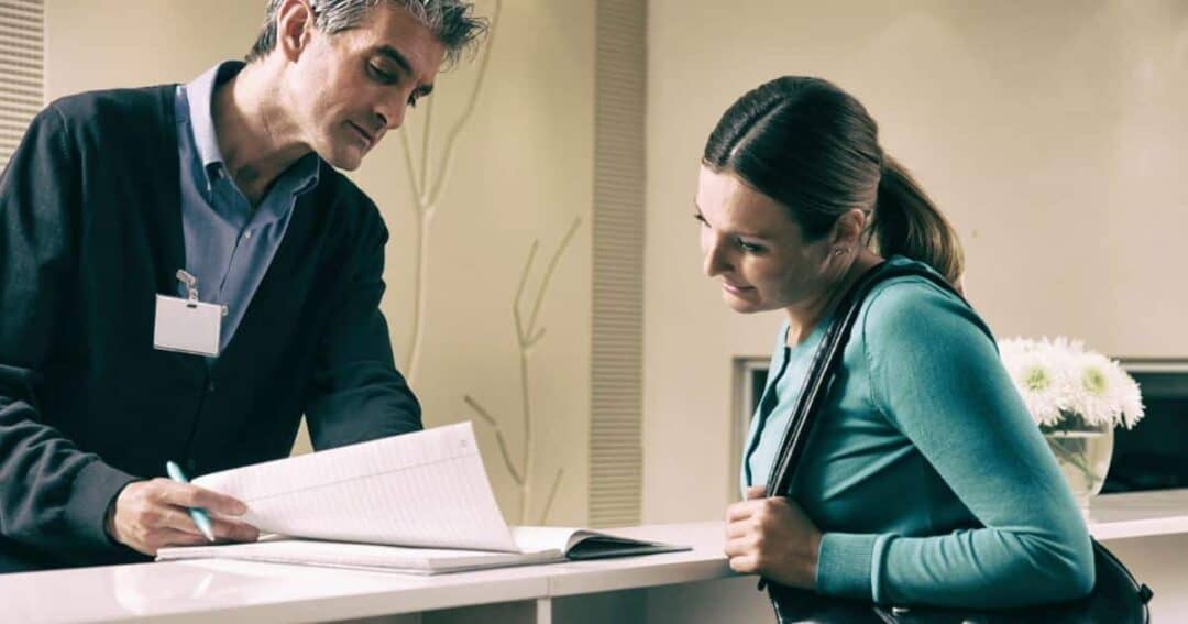 How can Patients and Caregivers Get Involved in Research? 1 A man in a cardigan and ID badge assists a woman in a teal hoodie at a reception desk. He points at a document while she leans over, observing. A vase with white flowers is on the counter.