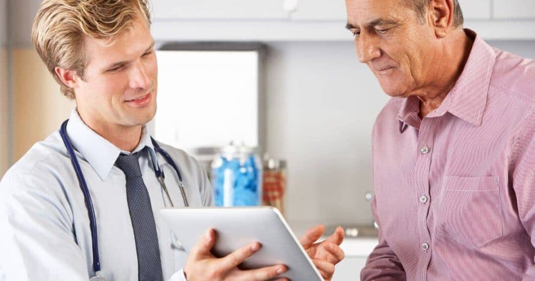 How can Patients and Caregivers Get Involved in Research? 2 A doctor with a stethoscope around his neck shows a tablet to an older man in a pink shirt. They are in a well-lit room, possibly a clinic, and are engaged in a discussion.