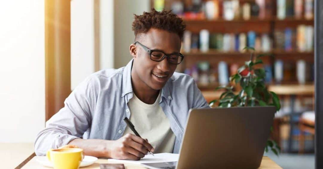 How can Patients and Caregivers Get Involved in Research? 6 A person wearing glasses sits patiently at a desk, diligently writing in a notebook while looking at a laptop. There's a cup of coffee and a smartphone on the table, suggesting they're deep into their research. The background is adorned with bookshelves and a potted plant.