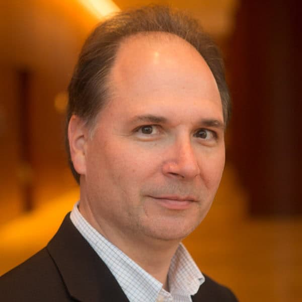 A man in a suit and collared shirt is looking at the camera. The background is softly blurred and warmly lit.