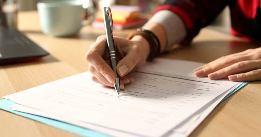 A person is filling out paperwork with a pen on a wooden desk. There's a cup and an open laptop nearby. The person is wearing a bracelet and a red and black plaid shirt. The scene suggests a workspace or office environment.