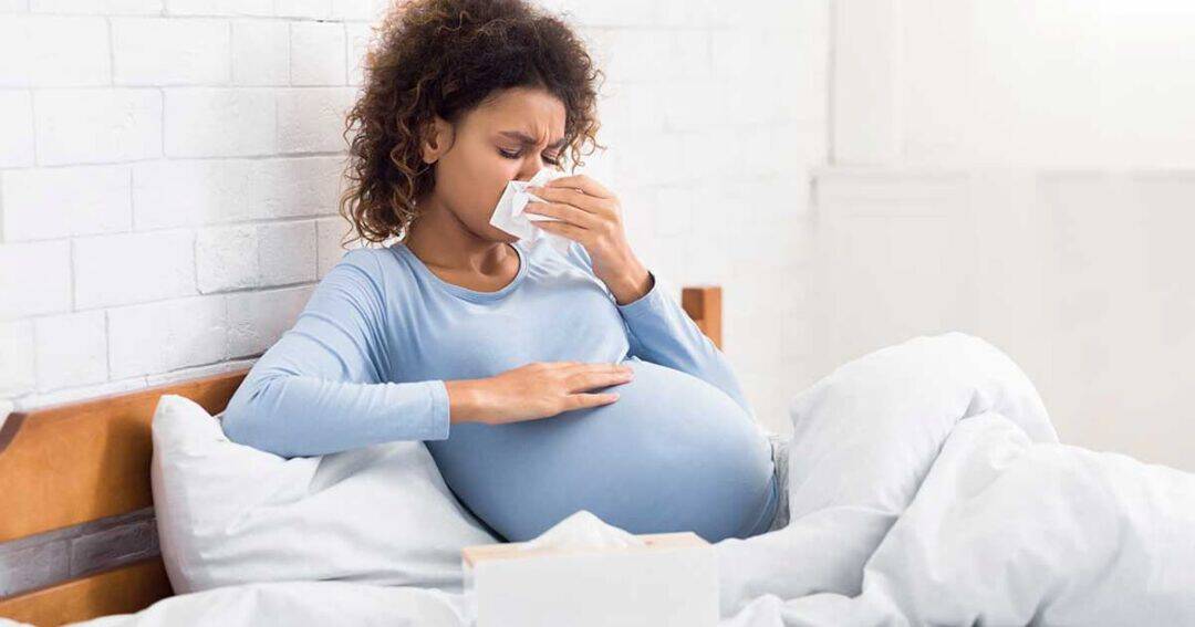 Pregnant woman sitting in bed, wearing a blue shirt, blowing her nose with a tissue, and touching her stomach. She looks unwell, with a tissue box nearby. The bed has white sheets and a light, airy atmosphere.