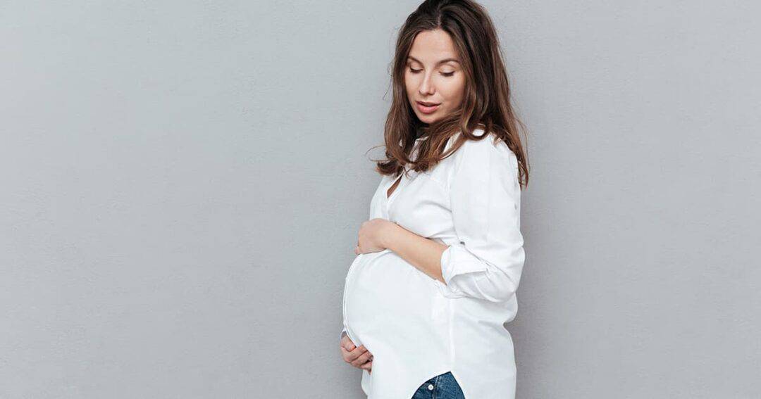 Asthma and Pregnancy 1 A pregnant woman stands sideways against a gray background, gently holding her belly. She is wearing a white shirt and blue jeans, looking down with a serene expression.