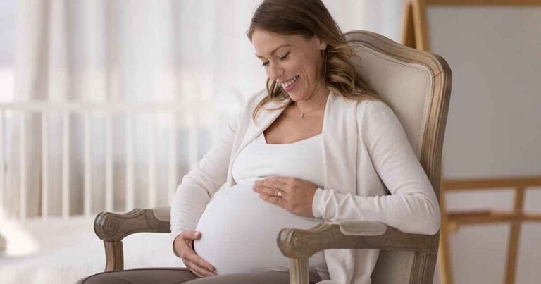 Your Trusted Resource 20 A pregnant woman sits in a cushioned chair, gently cradling her belly with both hands. She is smiling and looking down, wearing a white top and a light-colored cardigan. The background is softly lit and blurred, suggesting a calm, serene setting.