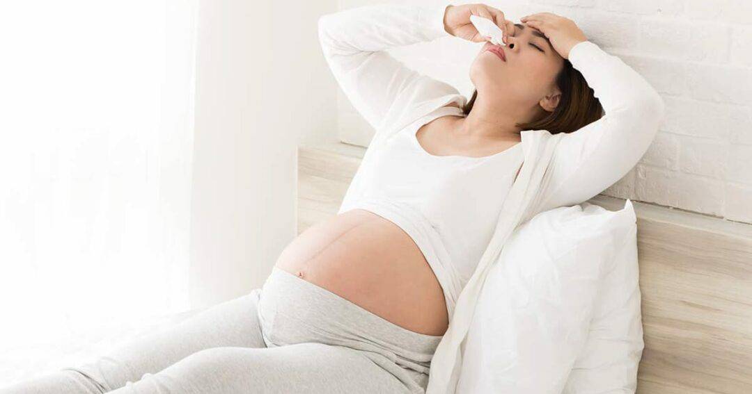 A pregnant woman sits on a bed, wearing light-colored lounge clothes. She holds her head with one hand and pinches her nose with the other, appearing to have a headache or discomfort. Pillows are visible in the background.