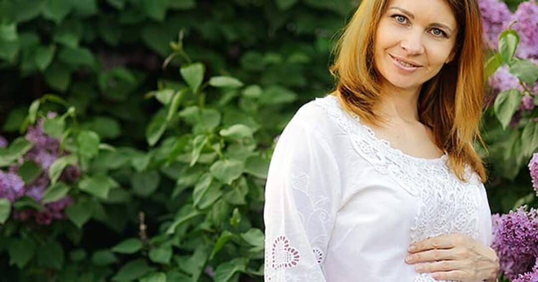 A woman with long brown hair is wearing a white dress and standing in front of a lush green background with purple flowers. She is smiling and gently resting one hand on her abdomen.