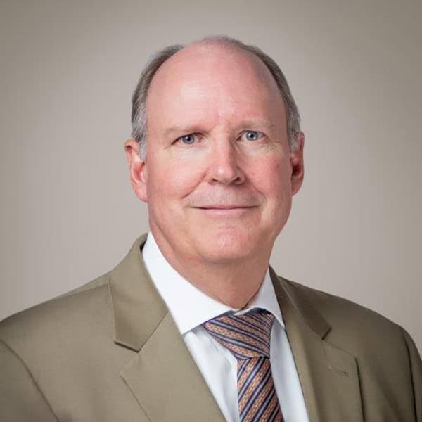 A middle-aged man wearing a tan suit jacket, white shirt, and patterned tie poses against a plain background. He has short gray hair and is smiling softly at the camera.
