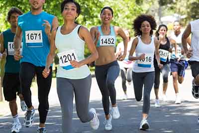 Ways to Give 5 A group of people running in a race, wearing numbered bibs. They are outdoors on a sunny day, surrounded by greenery. The runners appear focused and determined.