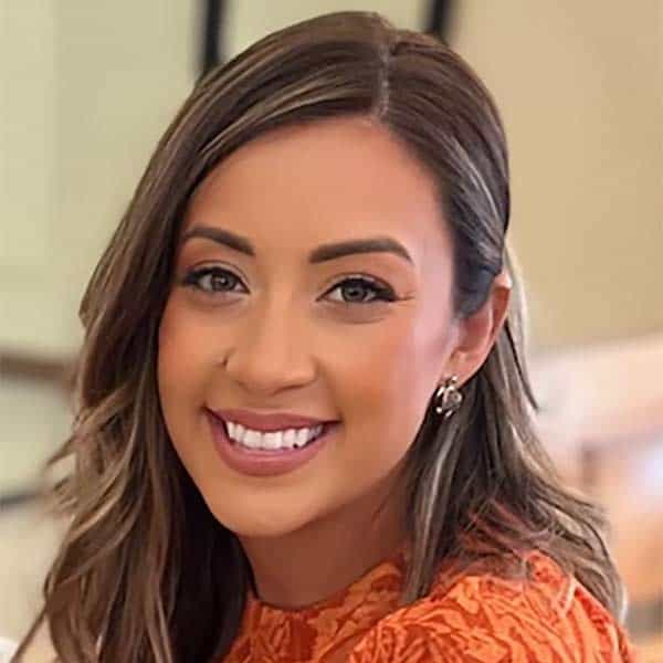 Smiling woman with long, light brown hair wearing an orange patterned top, posing indoors.