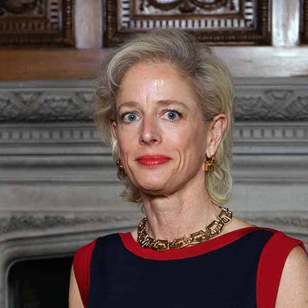 A woman with short blond hair stands in front of an ornate wooden and stone wall. She is wearing a sleeveless navy blue dress with red trim and gold earrings, with a matching necklace. She looks directly at the camera with a neutral expression.