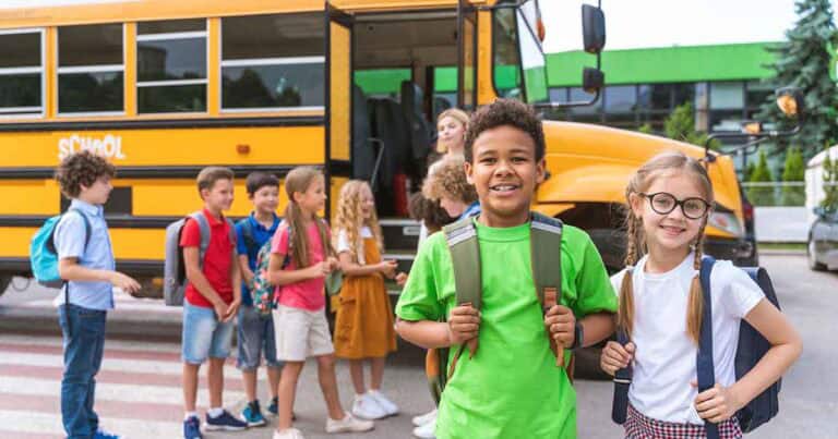 Your Trusted Resource 18 A group of smiling children with backpacks stand near a yellow school bus. Two kids, a boy in a green shirt and a girl in glasses and pigtails, stand in the foreground, while others chat and wait by the bus.