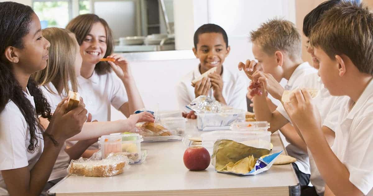 Planning for School with Food Allergies 1 A group of schoolchildren in uniforms sit around a table, enjoying lunch together. They have sandwiches, snacks, and an apple. The atmosphere is cheerful and friendly.