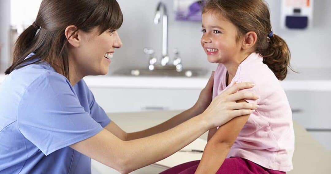 A smiling nurse in scrubs kneels beside a young girl sitting on an exam table. The nurse gently holds the girl's arms, and they both seem cheerful. A sink and medical equipment are visible in the background, indicating a doctor's office.