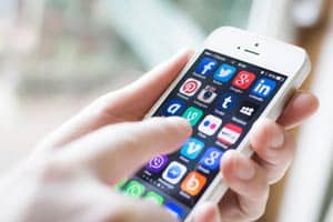 A person holding a smartphone displaying a variety of social media and communication apps, including Facebook, Twitter, Instagram, LinkedIn, and WhatsApp, among others. The person's hand is actively interacting with the screen.