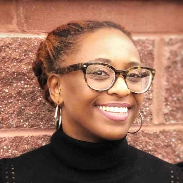 Black People Like Me Virtual Conference Series 2 Smiling person with tortoiseshell glasses and hoop earrings stands in front of a brick wall, wearing a black turtleneck. Hair is styled in an updo.