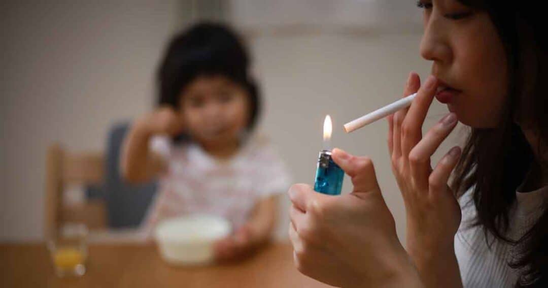 A woman holds a lit lighter to a cigarette in her mouth. In the background, a child sits at a table with a bowl and a glass of orange juice, appearing out of focus. The scene suggests an indoor setting.