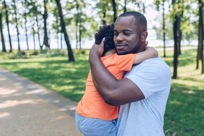 Ways to Give 6 A man gently holds a small child in his arms while standing on a park pathway. The child wears an orange shirt, and they are surrounded by green trees and grass, creating a serene and comforting atmosphere.