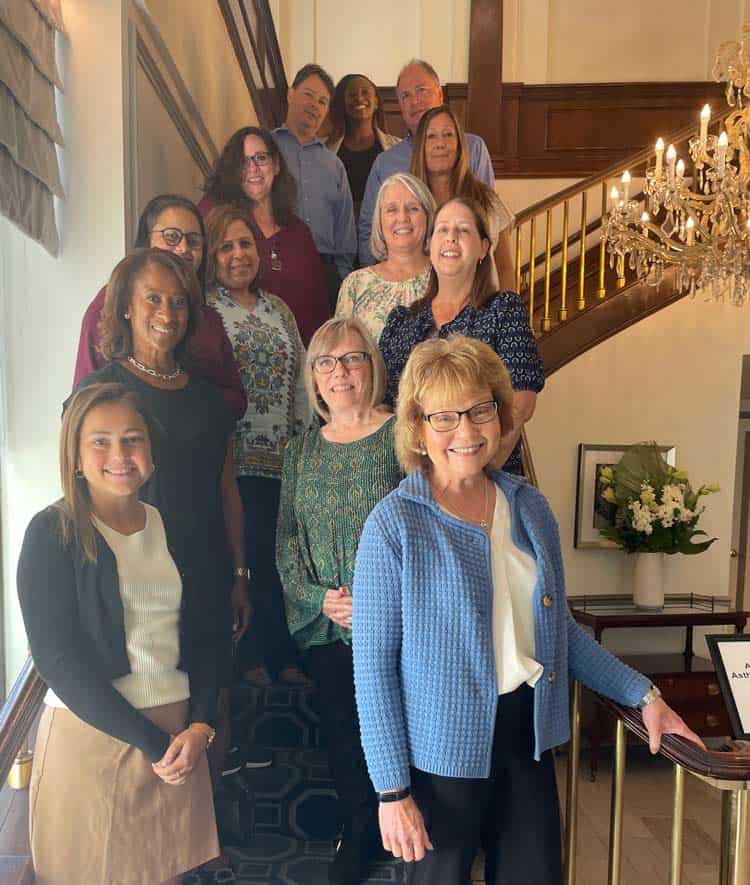 A group of thirteen people standing on a staircase, smiling at the camera. They are inside a well-lit room with a chandelier and some decorative plants. The atmosphere appears friendly and professional.