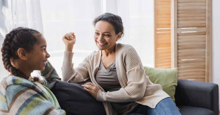 A woman in a beige cardigan sits on a couch, smiling and talking to a young girl with braided hair who is wrapped in a striped blanket. They are in a bright room with sunlight streaming through the window.