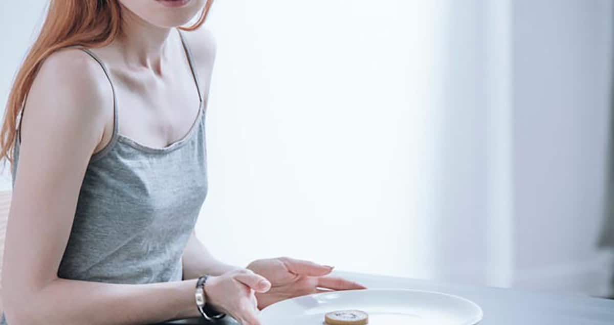 A person with long red hair, wearing a gray tank top, is sitting at a table. They have an empty white plate in front of them with a small pastry on it, reflecting their food anxiety. The background is softly lit with a white curtain.