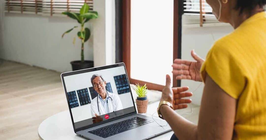 A woman in a yellow shirt is having a video call with a doctor on her laptop. The laptop is placed on a white table with a small potted plant. The room has large windows and light-colored flooring.
