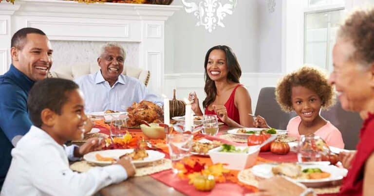 A diverse family of six enjoys a festive meal at a dining table decorated with autumn leaves and small pumpkins. Everyone is smiling and talking, contributing to a warm and cheerful atmosphere.