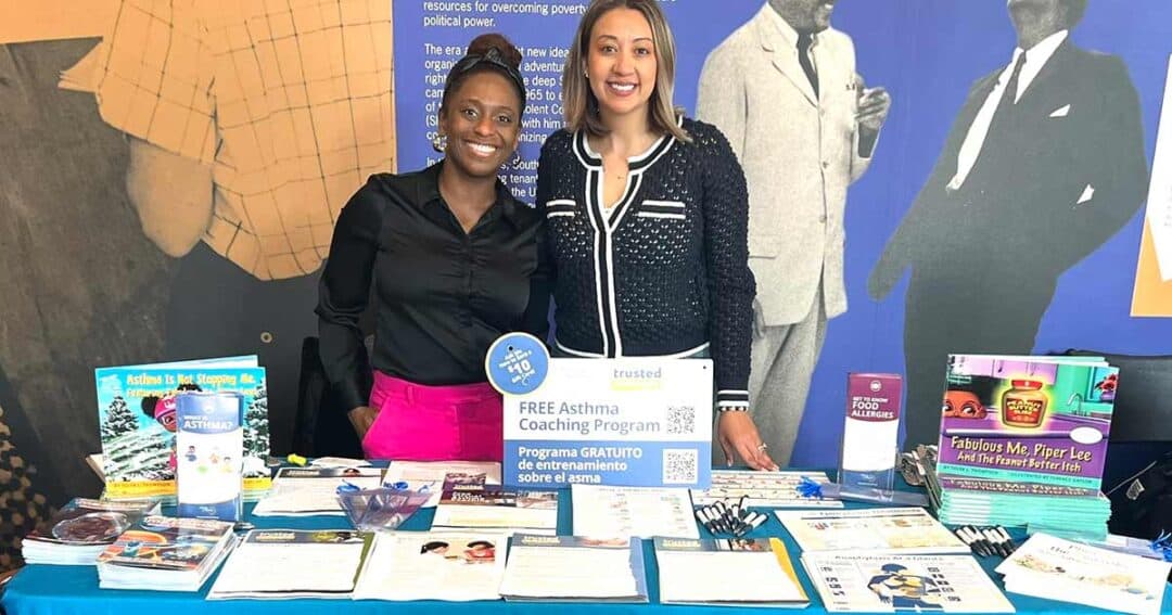 Two women stand behind a table with pamphlets and booklets about asthma coaching and health resources. A sign reads &ldquo;Free Asthma Coaching Program.&rdquo; The background features images of historical figures.