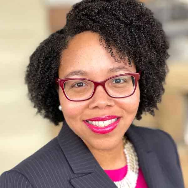 A woman with curly black hair wearing red glasses, a dark blazer, and a pink top smiles at the camera. She is also wearing a pearl necklace and earrings. The background is blurred and neutral.