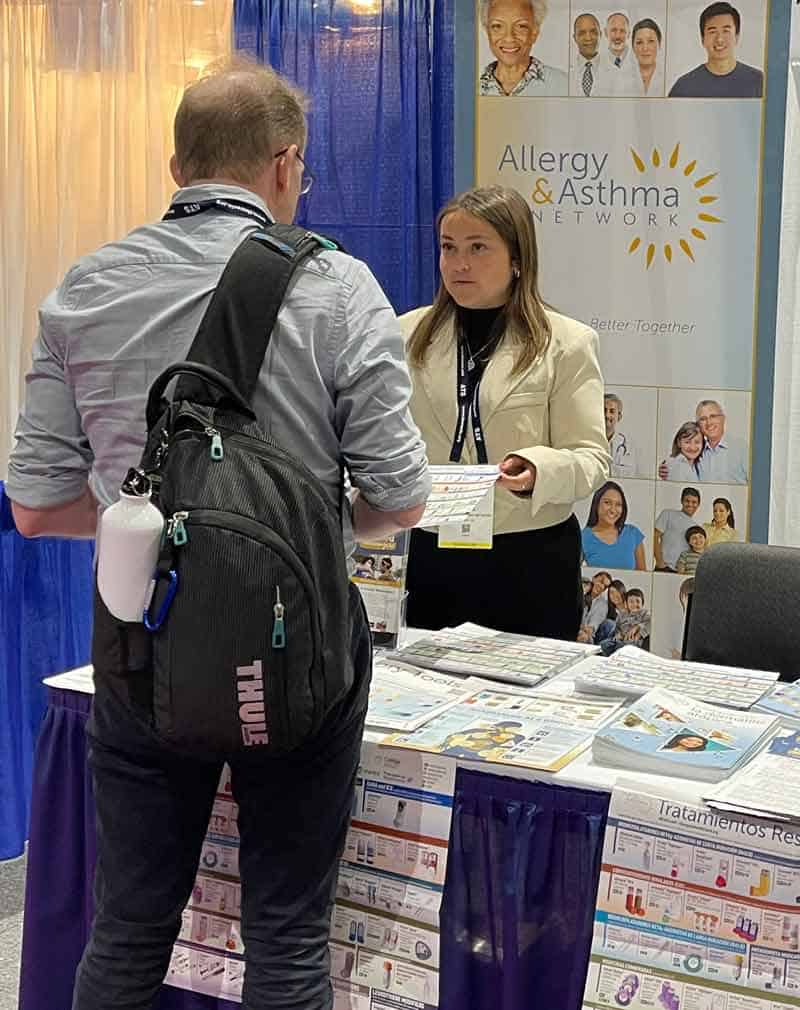A woman at a booth for the Allergy & Asthma Network is speaking with a man. She is holding a brochure, and informational materials are spread on the table. The backdrop displays the network's logo and images of diverse individuals.