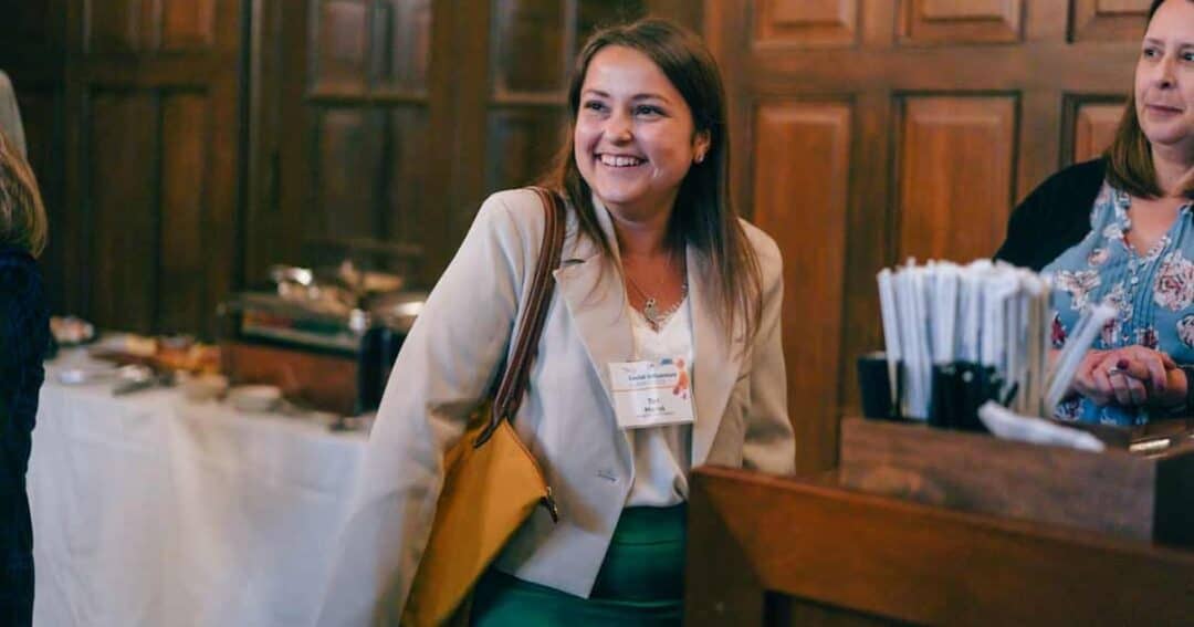 A woman smiling while wearing a beige blazer and holding a yellow bag, stands in a room with wood-paneled walls. She has a name tag on her blazer. Another woman, partially visible, is standing nearby. In the background are tables with covered dishes.