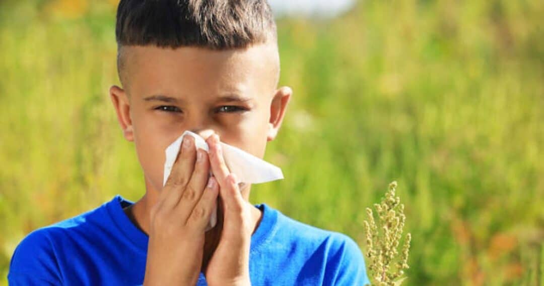 A young boy in a blue shirt stands outdoors, holding a tissue to his nose. He appears to be sneezing or has a runny nose, likely due to allergies. The background features green foliage and grass.