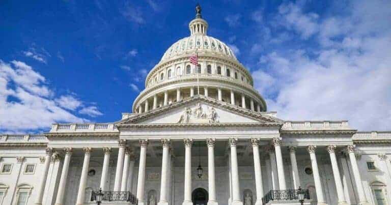 The image shows the United States Capitol building with its iconic dome and columns, under a partly cloudy blue sky. An American flag waves on top of the structure, highlighting the building's grandeur and historical significance.
