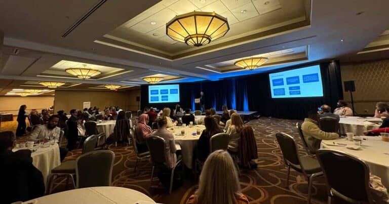 A conference room filled with attendees seated at round tables. A presenter stands on a stage with a large screen displaying slides. The room is lit with warm overhead lights, creating an engaging atmosphere for the audience.