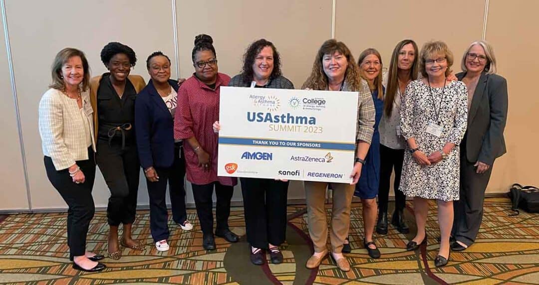 A group of ten people stand indoors, smiling, and holding a sign that reads "USAsthma Summit 2023." The sign displays logos for sponsors AMGEN, Sanofi, AstraZeneca, and Regeneron. They are dressed in business casual attire.