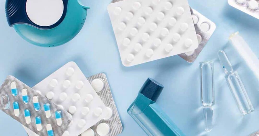 Various blister packs of pills, ampoules, an inhaler, and a pill organizer arranged on a light blue background. The packs contain white and blue capsules, and transparent glass ampoules are scattered around.