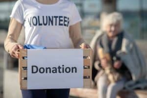 A person wearing a "VOLUNTEER" shirt holds a donation box filled with items. In the background, an elderly person sits on a bench, wrapped in a blanket. The scene conveys a charitable and caring atmosphere.