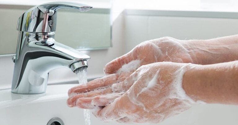 A person washing hands under a running faucet with soap lathered. The chrome faucet is part of a white sink in a bathroom setting.