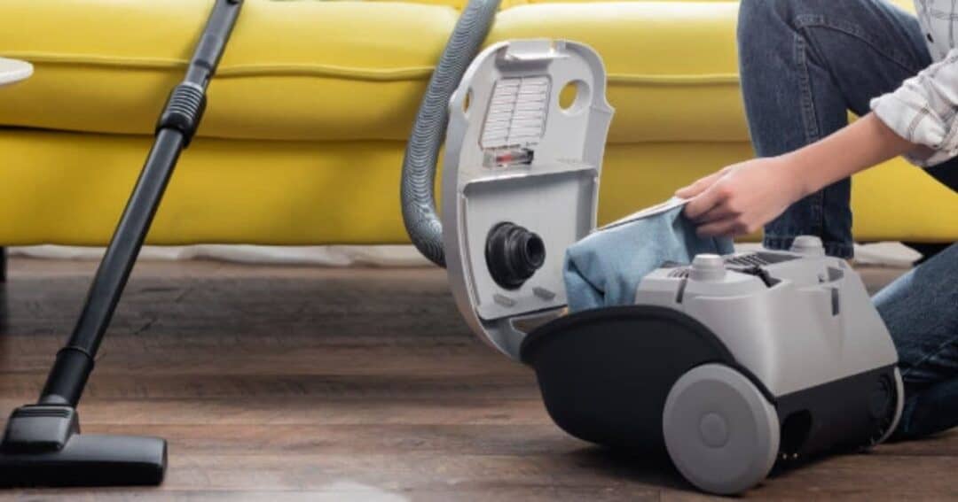 A person is kneeling on the floor, replacing the bag in a gray canister vacuum cleaner. The vacuum is on a wooden floor next to a yellow sofa. A vacuum head with hose is extended across the floor.