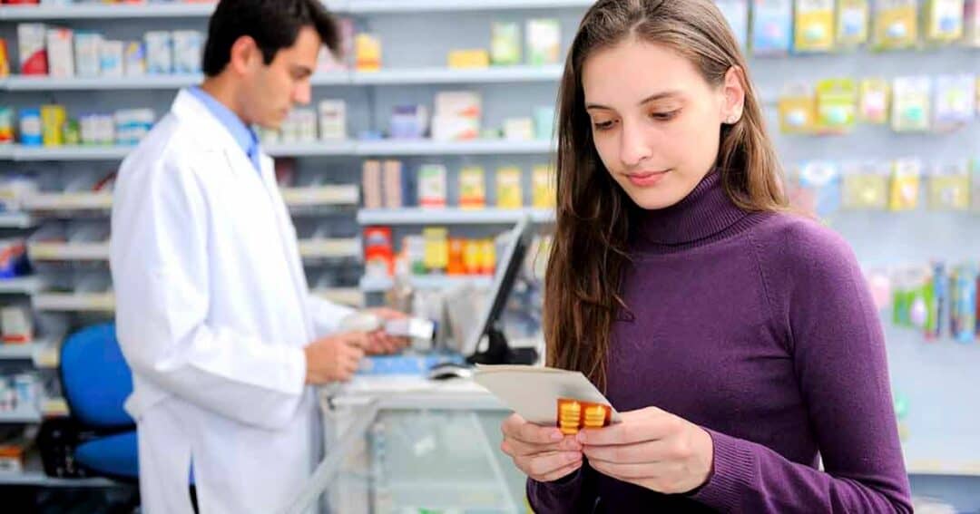 A woman in a purple sweater holds medication and a prescription while standing in a pharmacy. A pharmacist in a white coat is working in the background, surrounded by shelves with various health products.